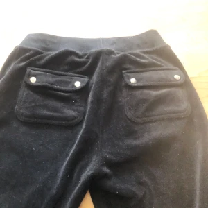 Svarta velourbyxor Juicy Couture XS - Svarta mjukisbyxor från Juicy Couture i velour med hög midja och raka ben. Byxorna har två fickor bak med lock och silverfärgade knappar. Perfekta för en chill och trendig look.