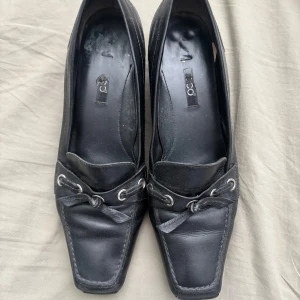 Svarta loafers med klack från Ecco - Klassiska svarta loafers från Ecco i skinn med fyrkantig tå, låg klack och dekorativ snörning framtill. Snygga detaljer med metallöljetter och rosett. Perfekta för dig som gillar en stilren och tidlös look.