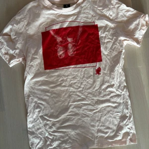 T-shirt - Vit t-shirt i bomull med en stor röd grafisk print på bröstet som föreställer ett par händer som knyter skosnören. Klassisk passform och rund halsringning. Perfekt för dig som gillar streetstyle och unika tryck.