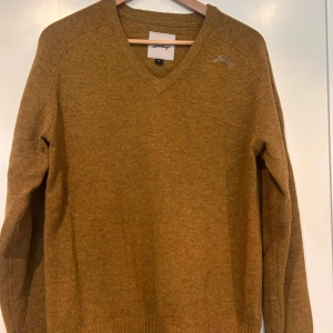 ”Senapsgul” stickad ulltröja från Lundhags - Säljer en ”senapsgul” stickad ulltröja från Lundhags i storlek M. Tröjan har v-ringning, långa ärmar och broderad logga på bröstet. Perfekt för dig som gillar en klassisk och enkel stil med lite outdoor-vibe.