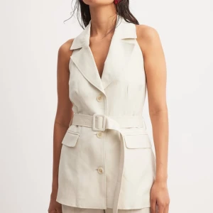 Beige omlottväst med bälte - Snygg beige väst i omlottmodell med bred krage, knappar framtill och matchande bälte i midjan. Västen är ärmlös och har öppen rygg med resår, samt två fickor med lock framtill. Perfekt för en trendig och stilren look. Väldigt väldigt fin men jag får bara ingen användning för den, legat i garderoben i evigheter men aldrig använd nypris 699