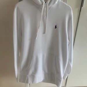 Vit hoodie från Ralph Lauren - Klassisk vit hoodie från Ralph Lauren med blå broderad logga på bröstet. Tröjan har huva med snörning, känguruficka . Några märken på arm därav billigt pris. 