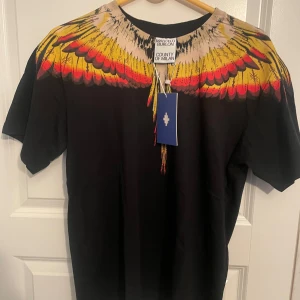 Svart t-shirt Marcelo Burlon med fjädertryck - Svart t-shirt från Marcelo Burlon County of Milan med ikoniskt färgglatt fjädertryck i gult och rött över axlar och halsringning. Klassisk passform och korta ärmar, tillverkad i mjuk bomull. Perfekt statement-plagg för dig som gillar streetwear.