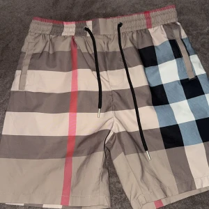 Burberry rutiga shorts med snörning - Snygga Burberry shorts med klassiskt rutigt mönster i beige, svart, blått, vitt och rött. De har elastisk midja med svart snörning och praktiska fickor. Perfekta för sommaren och ger en lyxig vibe till din outfit. Priset kan diskuteras vid snabbt affär.