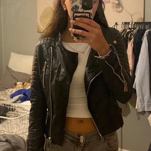 Svart skinnjacka med dragkedjor - Snygg svart skinnjacka med coola detaljer som dragkedjor framtill och på ärmarna, quiltade partier på axlar och armar samt spännen i midjan. Jackan har en figurnära passform. Storlek 36 men känd mer som en 34 