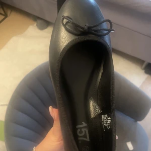 Svarta ballerinaskor med rosett - Klassiska svarta ballerinaskor från 157 med rund tå och en söt liten rosett framtill. Skorna är gjorda i syntetmaterial med en platt sula, perfekta för en clean och stilren look.