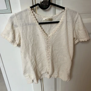 Vit blus med snörning från Zarella - Supersöt vit blus från Zarella med korta ärmar och v-ringning. Blusen har dekorativ snörning framtill och fina virkade kanter längs halsringning och ärmslut. Materialet är strukturerat och känns mjukt mot huden. Perfekt för dig som gillar romantiska detaljer!