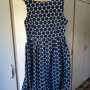 Marinblå prickig ärmlös klänning - Snygg ärmlös klänning med marinblå stora prickar på vit botten. Klänningen har en markerad midja och utställd kjol som ger en klassisk siluett. Perfekt för dig som gillar retrovibbar och vill sticka ut med ett coolt mönster.