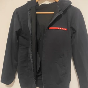 Tracksuit från Prada - Svarta mjukisbyxor från Prada med snyggt rött logoband på benet. Byxorna har dragsko i midjan, två sidofickor och en avslappnad passform. Materialet är mjukt och skönt, perfekt för chill dagar. Stilren design med exklusiv känsla. Ingen dragkedja på jackan.