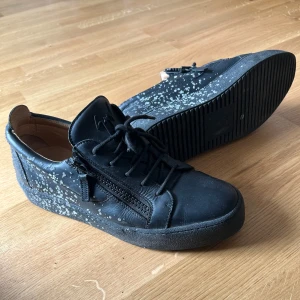 giuseppe skor - Säljer ett par svarta sneakers från Giuseppe Zanotti med coolt vitt stänkmönster på sidorna och sulan. Skorna har snörning, dragkedja på sidan och är gjorda i skinn med en platt sula. Perfekt för dig som gillar streetstyle och vill sticka ut med detaljer. Använda en sommar. Finns tyvärr ingen box