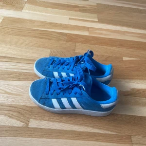 Blå Adidas Campus sneakers, stl 38⅔ - Säljer ett par blå Adidas Campus sneakers med klassiska vita ränder på sidan och vit sula. Skorna har snörning och är gjorda i mocka med rund tå. Adidas-logga på hälen och Campus-tryck på sidan. Perfekt för dig som gillar retrostil och streetwear. Dem har tecken på användning men inget som man tänker på mycket. 