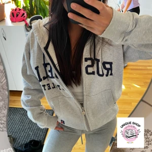Grå zip-hoodie från Russell Athletic - Grå hoodie med dragkedja och mörkblå Russell Athletic-tryck över bröstet. Jackan har huva med snörning, två fickor fram och ribbade muddar. Perfekt för chill eller sportig stil. Snyggt loose fit och mjukt bomullsmaterial.