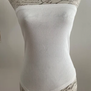 Vit ribbad bandeau tubtopp - Superenkel och fräsch vit bandeau tubtopp med ribbad struktur. Sitter tight och har en rak, axelbandslös design som är perfekt för varma dagar. Materialet är mjukt och stretchigt, vilket gör den bekväm att bära hela dagen. Passar till jeans, kjol eller shorts.