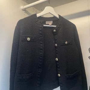 Svart cardigan jacka från H&M XS - Svart stickad cardigan jacka från H&M i storlek XS. Jackan har dekorativa silvriga knappar framtill och på fickorna, samt en snygg kantdetalj längs öppningen. Perfekt för dig som gillar klassisk och stilren look med en twist.