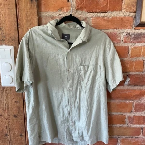 Ljusgrön kortärmad skjorta från H&M - Snygg ljusgrön skjorta från H&M i regular fit. Skjortan har korta ärmar, klassisk krage, knäppning framtill och en bröstficka. Materialet är mjukt och luftigt linne, perfekt för varma dagar. Enkel och stilren design som funkar till många olika looks.