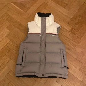 Peak Performance dunväst grå/vit - Säljer en dunväst från Peak Performance i grått och vitt med hög krage och broderad logga på bröstet. Västen har dragkedja framtill, justerbar nederkant och röda detaljer vid axlarna. Perfekt för lager-på-lager under kyliga dagar.
