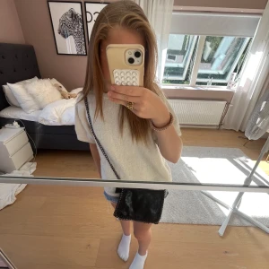 Svart axelväska med kedjedetaljer - Snygg svart axelväska med kedja som axelrem och coola kedjedetaljer runt kanterna. Väskan har en fyrkantig form och är gjord i mocka, vilket ger en mjuk och lyxig känsla. Perfekt storlek för mobil, plånbok och andra essentials.