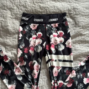 Blommiga leggings från Stronger - Snygga svarta leggings från Stronger med blommigt mönster i rosa, vitt och rött. Bred midja med logga och vita ränder på ena benet. 