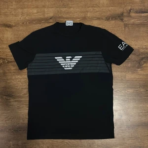 Svart t-shirt från Emporio Armani - Snygg svart t-shirt från Emporio Armani med det ikoniska EA7-logotypen tryckt på bröstet och ärmen. T-shirten har en stilren design med horisontella ränder över bröstet. Perfekt för en casual look.