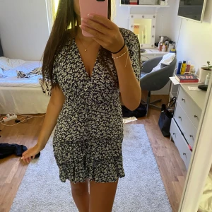 Blommig kortklänning med volang - Supersöt kortklänning i mörkblått med vitt blommönster. Klänningen har v-ringning, omlottliv och volangkant nedtill. Korta ärmar i luftig modell och knyt i midjan för snygg passform. Perfekt för sommarens alla tillfällen!