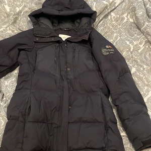 Svart Sail Racing dunjacka XS - Säljer en svart dunjacka från Sail Racing i storlek XS. Jackan har huva, flera dragkedjor framtill och snygg patch på ärmen med Sail Antarctica Expedition. Perfekt för kalla dagar och har en stilren, lite längre passform.