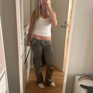Grå bootcut jeans med slitna knän - Säljer ett par grå jeans med låg midja och bootcut passform. De har slitna detaljer och hål vid båda knäna för en edgy vibe. Jeansen har klassiska fem fickor och är gjorda i jeansmaterial. Perfekt för dig som gillar en avslappnad och cool stil.