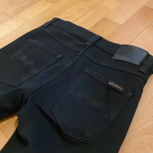 Svarta  jeans från Nudie Jeans - Säljer ett par svarta jeans från Nudie Jeans, tillverkade i 100% ekologisk bomull. Jeansen har klassiska fem fickor, svarta sömmar och Nudie Jeans logga på bakfickan. Snygg, smal passform som sitter nice längs benen och midja med knapp och dragkedja.