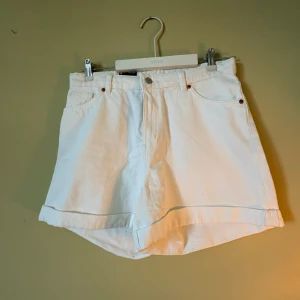 Vita högmidjade shorts från Monki - Snygga vita shorts från Monki med hög midja och klassisk femficksdesign. Tillverkade i ekologisk bomull med en loose passform och uppvikta benslut. Perfekta för varma dagar och en clean look. Bälteshällor och knappstängning framtill.