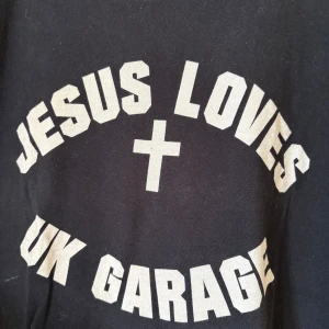 Svart Jesus Loves UK Garage t-shirt L - Svart t-shirt från Hot Haus Recs i storlek L med vit tryckt text 'Jesus Loves UK Garage' och ett kors på framsidan. Baksidan har ett graffiti-inspirerat tryck med Hot Haus Recs-loggan. Klassisk rund hals och korta ärmar, tillverkad i mjuk bomull.