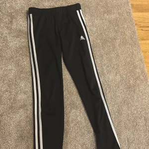 Svarta Adidas träningsbyxor 152 - Svarta träningsbyxor från Adidas med klassiska vita ränder längs sidorna och Adidas-logga på låret. Byxorna har elastisk midja och är gjorda i Primegreen polyester, vilket gör dem både sköna och hållbara. Perfekta för sport eller chill.