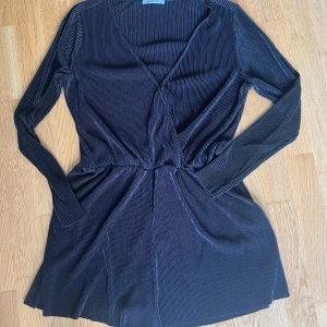 Svart jumpsuit från Gina Tricot - Snygg svart jumpsuit från Gina Tricot med omlott-design och v-ringning. Jumpsuiten har långa ärmar och är gjord i ett ribbat, mjukt material med mycket stretch.