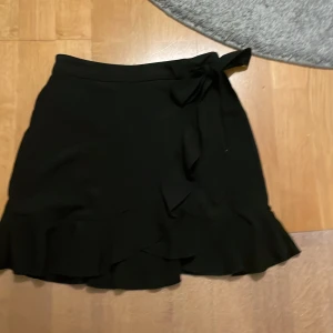 Svart omlottkjol med volang - Snygg svart kortkjol med omlott-design och volangkant. Kjolen har en knytning i midjan som ger en extra detalj och sitter bekvämt. Perfekt för dig som gillar stilrena plagg med lite edge. Materialet känns mjukt och följsamt.