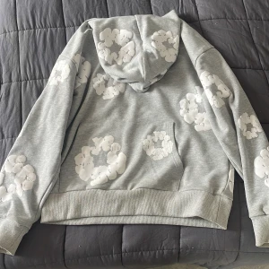 Grå hoodie med vita blommor Denim Tears - Säljer en grå hoodie från Denim Tears i storlek S med unika vita blommönster över hela tröjan. Tröjan har huva, magficka och ribbade muddar. Perfekt för dig som vill sticka ut med en cool streetwear-look.