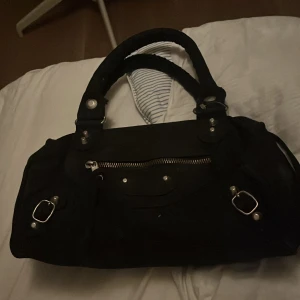 Svart handväska med silvriga detaljer - Cool svart handväska med två rejäla handtag och silvriga metalldetaljer. Väskan har en dragkedja framtill, spännen och nitar som ger en edgy vibe. Materialet ser ut att vara skinn och insidan har flera fack för dina grejer. Perfekt storlek för att få plats med allt du behöver.