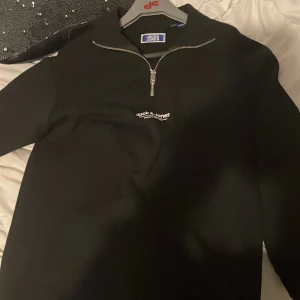 Svart half zip tröja Jack & Jones - Svart långärmad tröja från Jack & Jones med half zip-dragkedja och liten vit logga på bröstet. Tröjan har en clean look och är gjord i mjukt material som känns skönt mot huden. Perfekt för chill dagar och enkel att matcha med jeans eller joggers.