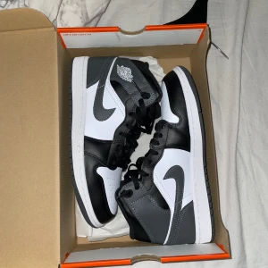 Nike Air Jordan 1 Mid svart/grå - Nike Air Jordan 1 Mid i svart, grått och vitt med klassisk Jordan Wings-logga på sidan. Skorna har snörning, rund tå och platt sula. Ovandelen är i skinn och syntet, med snygga kontraster mellan färgerna. Perfekt för dig som gillar streetstyle och vill sticka ut med en ikonisk sneaker.