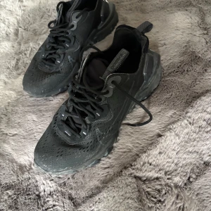 Nike React svarta sneakers - Nike react vision sneakers i helsvart med perforerad ovandel och chunky sula. Snyggt sportig design med snörning och diskret Swoosh på sidan. Ovandelen är i syntetmaterial och mesh för bra ventilation. 
