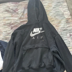 Svart Nike Air hoodie med huva - Svart hoodie från Nike Air med stor vit logga på bröstet. Tröjan har huva med snörning och en stor magficka framtill. Perfekt för chill eller streetwear, riktigt skön och stilren design. Passar dig som gillar sportiga och avslappnade outfits. Storlek s/m