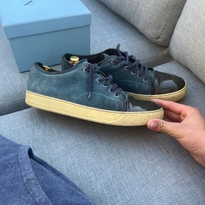 Lanvin blå sneakers med lackad tå - Snygga sneakers från Lanvin i mörkblå mocka med svart lackad tå och beige sula. Skorna har rund tå, platt sula och klassisk snörning. Perfekt för dig som gillar exklusiva detaljer och vill sticka ut med en unik kombo av material och färg. Kommer med og lådan! 