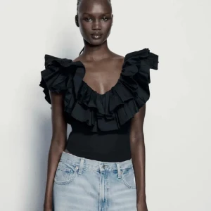 Svart volang body med v-ringning - Säljer en svart bodysuit med stor volang runt axlar och halsringning. Lappen är bortklippt då den var synlig. Har även i storlek M