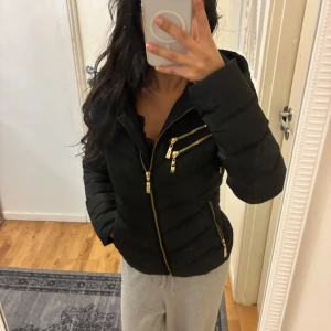 Svart pufferjacka från Hollies - INTRESSEKOLL! Svart pufferjacka från Hollies med guldiga dragkedjor och detaljer. Jackan har huva med snygg metallring och leopardmönstrat foder. Flera fickor med dragkedja framtill och quiltad design som ger en schysst form. 