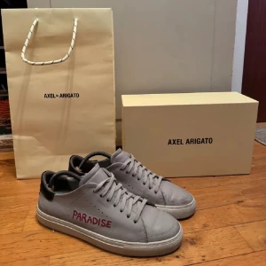 Grå Axel Arigato Paradise sneakers - Snygga grå sneakers från Axel Arigato med texten 'PARADISE' broderad i rött på sidan. Skorna har svart detalj på hälen, rund tå och platt sula. Tillverkade i skinn med klassisk snörning. Perfekta för dig som gillar stilrena och trendiga sneakers.