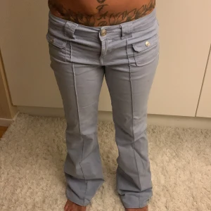 Ljusblå bootcut byxor med fickor - Snygga ljusblå bootcut byxor med framfickor med lock och silverfärgade knappar. Byxorna har hög midja och markerade sömmar längs benen för en clean look. Perfekta för dig som gillar en lite retro vibe och vill sticka ut med coola detaljer. Midjemått: 37cm
