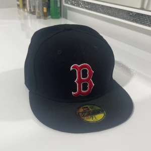 Svart Boston Red Sox keps New Era - Säljer en svart 59FIFTY keps från New Era med Boston Red Sox B-logga i rött och vitt framtill. MLB-logga broderad baktill. Kepsen är i klassisk flatbrim-modell och tillverkad i 100% polyester. Perfekt för dig som gillar sportig streetstyle.