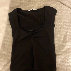 Svart topp med v-ringning från 157 - Svart topp från 157 i storlek S med snygg v-ringning och lätt rynkning framtill. Modellen är kortärmad och har en enkel, stilren look som funkar till många olika outfits. Mjuk bomullskänsla och figurnära passform.