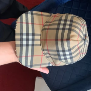 Burberry rutig keps med knäppning - Säljer en klassisk Burberry keps med ikoniskt rutigt mönster i beige, svart och rött. Kepsen har en böjd skärm och justerbar knäppning bak med svart läderrem. Märket Burberry London England syns tydligt baktill. Perfekt för dig som gillar street och designer.
