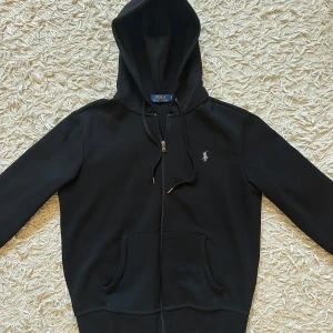 RALPH LAUREN- Svart  ziphoodie  - Säljer en klassisk hoodie från Polo Ralph Lauren med klassisk broderad logga på bröstet. Tröjan har huva med snörning, dragkedja framtill och två fickor. Materialet är mjuk bomull och passformen är normal, varken slim eller oversized. Passar dig från 172-183. Perfekt för chill dagar och streetwear. Den är helt ny- oanvänd och tagsen medföljer! 