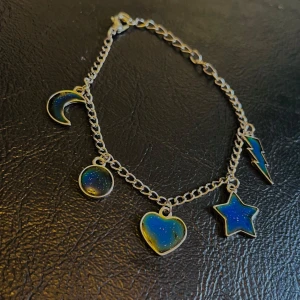 Silverfärgat armband med blå berlocker - Coolt silverfärgat armband med fem berlocker: måne, rund, hjärta, stjärna och blixt. Alla berlocker har en blå och grön glittrig finish som ger en galaktisk vibe. Perfekt för dig som gillar unika detaljer och vill sticka ut med din stil.