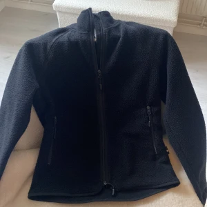 Svart fleecejacka från 157 XS - Svart fleecejacka från 157 i storlek XS. Jackan har hög krage, hel dragkedja framtill och två sidofickor med dragkedja. Materialet är mjuk fleece som håller dig varm och bekväm. Perfekt för kyliga dagar i höst.
