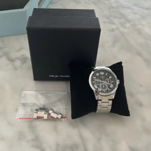 Silverfärgad klocka från Regal - Stilren klocka från Regal med silverfärgad länkarmband och svart urtavla. Urtavlan har romerska siffror och tre mindre dekorativa visare. Klockan levereras med extra länkar och originalask. Perfekt accessoar för en clean look.